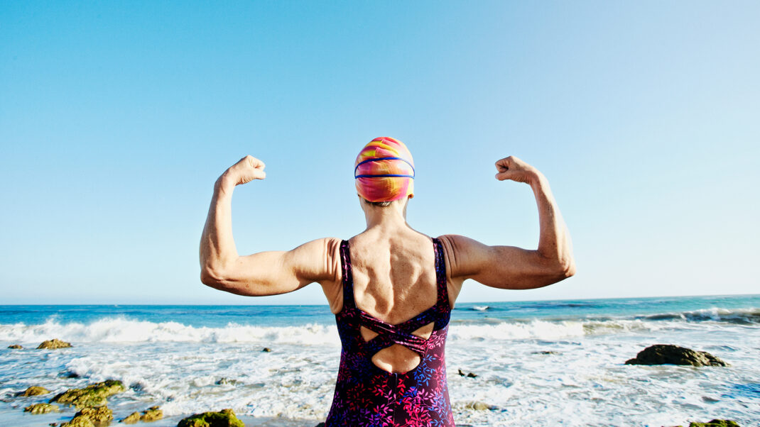 Older woman flexing biceps in front of ocean to show benefits of swimming for menopause symptoms