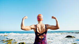 Older woman flexing biceps in front of ocean to show benefits of swimming for menopause symptoms